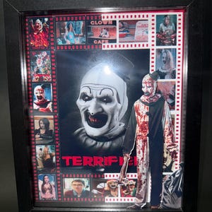 May include: Framed artwork with a horror movie theme. The central image is a close-up of a clown's face with the word "TERRIFIER" in red. Smaller images and film strip borders surround the main image, all within a black frame.