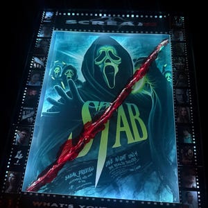 Könnte beinhalten: Ein rechteckiger Leuchtkasten mit einem Filmplakat für Stab, das eine Ghostface-Figur mit dunkler Kapuze und einen großen, diagonalen roten Schnitt in der Mitte zeigt. Der Rand enthält Filmstreifen und den Text What's your favorite scary movie?