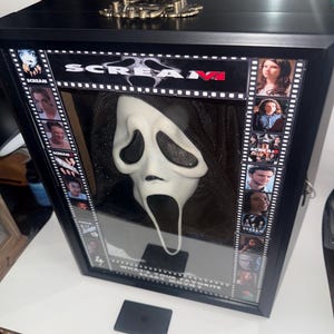 May include: A framed display featuring a Scream mask. The mask is white with black eye and mouth openings. The frame is black with the text "Scream VI" and film strip borders with character images. A black box is in front of the frame.