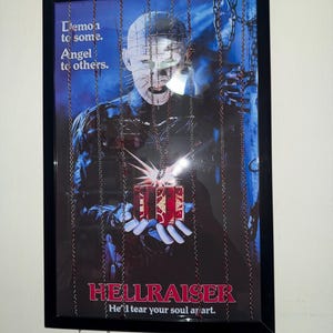 Puede incluir: Póster enmarcado de la película de terror Hellraiser. El póster presenta un personaje con alfileres en la cabeza sosteniendo una caja de regalo roja. El texto incluye "Demonio para algunos. Ángel para otros." y "Te destrozará el alma."