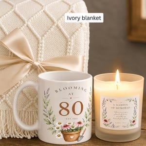 May include: An ivory knit blanket with a satin ribbon, a white ceramic mug featuring the text "BLOOMING AT 80 EST. 1945" with floral and garden tool graphics, and a lit candle in a frosted glass jar labeled "A GARDEN OF MEMORIES" with floral art.