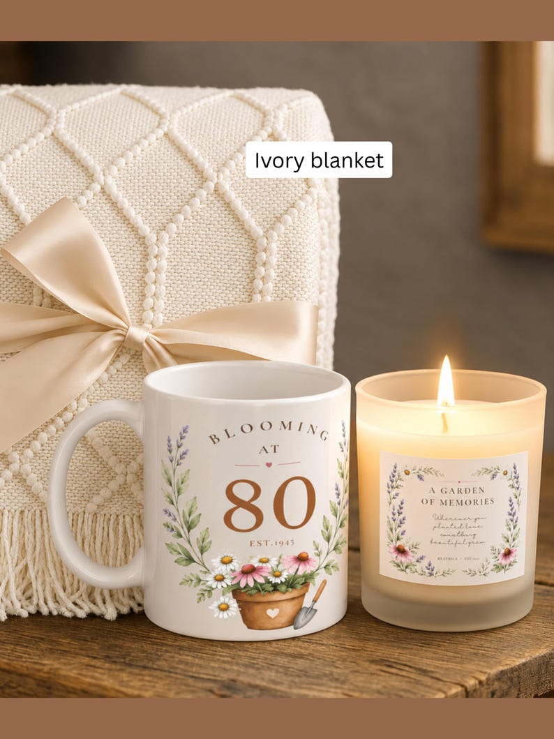 May include: An ivory knit blanket with a satin ribbon, a white ceramic mug featuring the text "BLOOMING AT 80 EST. 1945" with floral and garden tool graphics, and a lit candle in a frosted glass jar labeled "A GARDEN OF MEMORIES" with floral art.