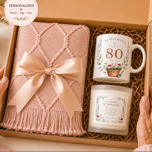 May include: A gift box containing a dusty pink knitted throw blanket with a satin ribbon, a white ceramic mug with the text "BLOOMING AT 80" and floral graphics, and a glass candle with a label reading "A GARDEN OF MEMORIES" and floral accents.