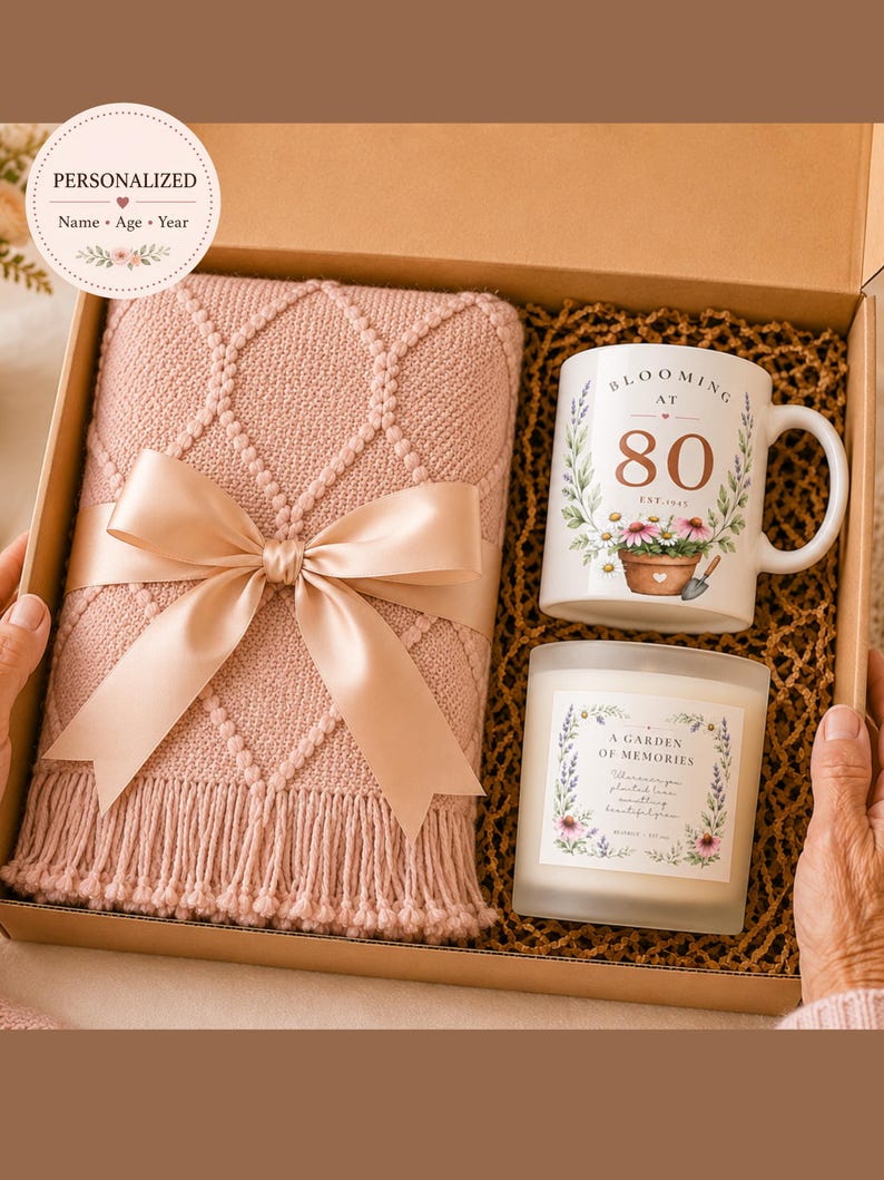 May include: A gift box containing a dusty pink knitted throw blanket with a satin ribbon, a white ceramic mug with the text "BLOOMING AT 80" and floral graphics, and a glass candle with a label reading "A GARDEN OF MEMORIES" and floral accents.