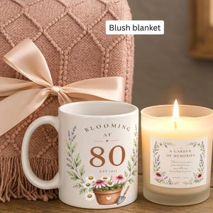 May include: A white ceramic mug featuring a floral design, the text "BLOOMING AT 80 EST. 1945," and a potted plant illustration, next to a lit candle in a frosted glass jar with a floral label, all placed before a folded blush blanket with a satin ribbon.