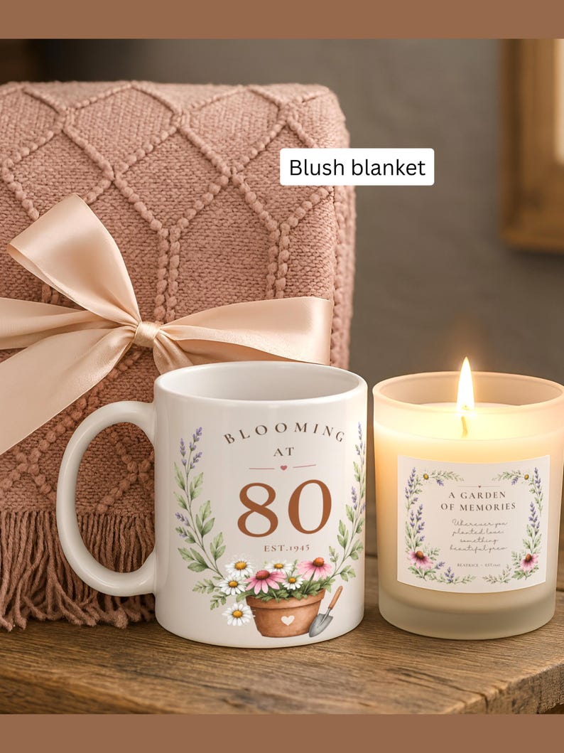 May include: A white ceramic mug featuring a floral design, the text "BLOOMING AT 80 EST. 1945," and a potted plant illustration, next to a lit candle in a frosted glass jar with a floral label, all placed before a folded blush blanket with a satin ribbon.