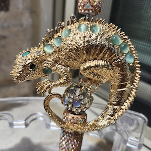 May include: A gold-colored pen featuring a detailed lizard design adorned with teal and clear gemstones. The pen's body is embellished with iridescent crystals, and the top has a small, gem-studded cap. The pen is a decorative writing instrument.