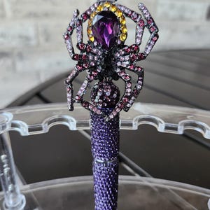 Puede incluir: Un bolígrafo con cuerpo incrustado de cristales morados y un adorno en forma de araña. La araña está decorada con pedrería rosa y morada, una gran piedra central morada en forma de lágrima y un halo de piedras amarillas.