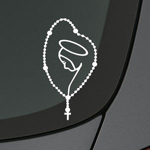 Może przedstawiać: Biała naklejka winylowa przedstawiająca minimalistyczny wzór różańca z sylwetką Matki Boskiej i aureolą. Różaniec zawiera krzyż na dole. Naklejka jest naklejona na ciemnoszarej powierzchni, prawdopodobnie szybie samochodu.