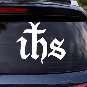 Può includere: Adesivo bianco IHS su un finestrino dell'auto. L'adesivo, in un carattere gotico, mostra le lettere IHS. Il simbolo religioso è apposto sul lunotto posteriore di un veicolo nero, offrendo un netto contrasto.