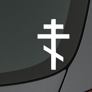 Op de afbeelding: Een witte sticker met een Oost-Orthodox kruis. Het heeft een horizontale balk bovenaan, een tweede balk eronder en een schuine balk onderaan. Op een donkergrijze ondergrond, vermoedelijk een voertuig.