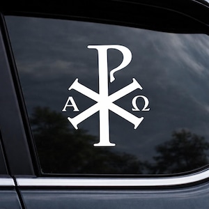 May include: White Chi Rho symbol car decal. The symbol is composed of the Greek letters Chi (X) and Rho (P), with the letters Alpha (A) and Omega (Ω) on either side. The decal is affixed to a car window, with a dark background.