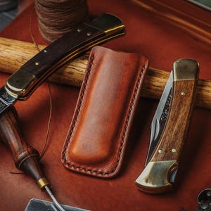 May include: Two folding pocket knives with wooden handles and brass bolsters, one featuring an etched blade, positioned alongside a tan leather sheath. The knives are displayed on a brown leather surface with a wooden tool handle nearby.