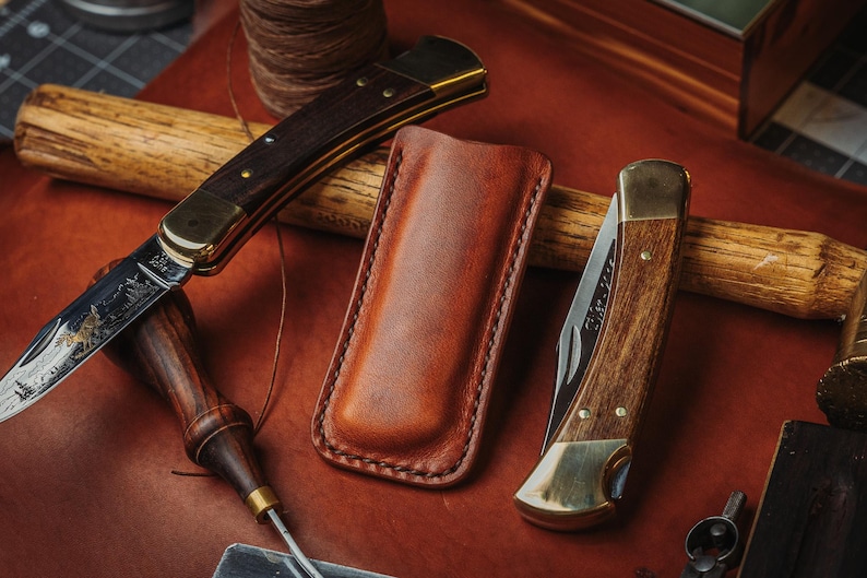 May include: Two folding pocket knives with wooden handles and brass bolsters, one featuring an etched blade, positioned alongside a tan leather sheath. The knives are displayed on a brown leather surface with a wooden tool handle nearby.