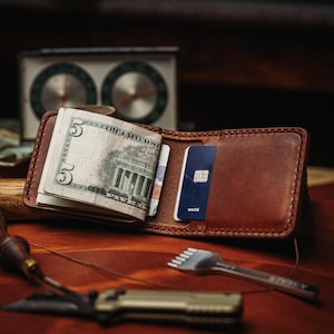 May include: Open brown leather wallet with cash and a blue credit card. A small knife and a metal tool are in the foreground. A vintage clock is in the background.