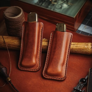 May include: Two tan leather knife sheaths with dark brown stitching, each containing a folding knife with a brass bolster and wooden handle. The sheaths are rectangular with rounded bottom corners and a molded shape to fit the knives securely.