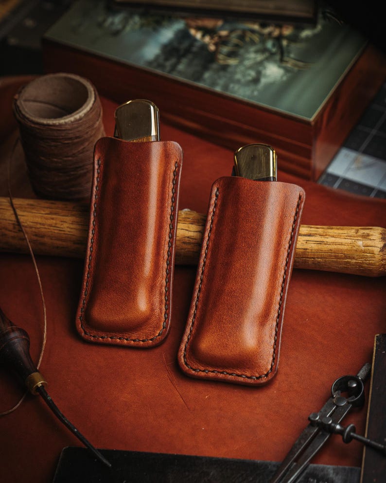 May include: Two tan leather knife sheaths with dark brown stitching, each containing a folding knife with a brass bolster and wooden handle. The sheaths are rectangular with rounded bottom corners and a molded shape to fit the knives securely.