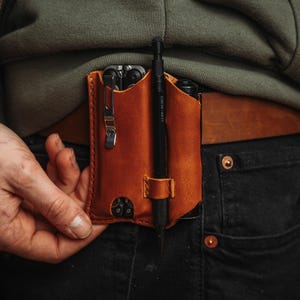 Może przedstawiać: Brązowa skórzana pochwa na multi-narzędzie z długopisem i multi-narzędziem w środku. Pochwa jest przymocowana do brązowego skórzanego paska. Osoba ma na sobie zieloną bluzę z kapturem i czarne dżinsy.