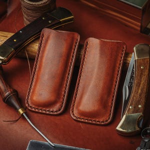 May include: Two rectangular brown leather knife sheaths with visible stitching along the edges, positioned between two folding pocket knives with wooden handles and brass bolsters, resting on a dark brown leather surface with crafting tools.