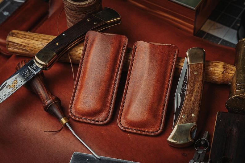 May include: Two rectangular brown leather knife sheaths with visible stitching along the edges, positioned between two folding pocket knives with wooden handles and brass bolsters, resting on a dark brown leather surface with crafting tools.
