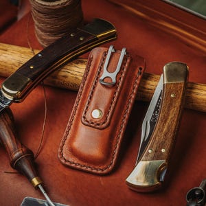 May include: Two folding pocket knives with wooden handles and brass bolsters, one open and one closed, alongside a tan leather sheath with a metal belt clip. The open knife blade features an etched landscape scene with trees and a mountain range.