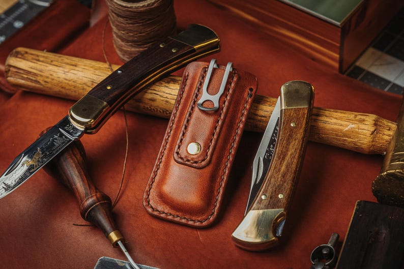 May include: Two folding pocket knives with wooden handles and brass bolsters, one open and one closed, alongside a tan leather sheath with a metal belt clip. The open knife blade features an etched landscape scene with trees and a mountain range.
