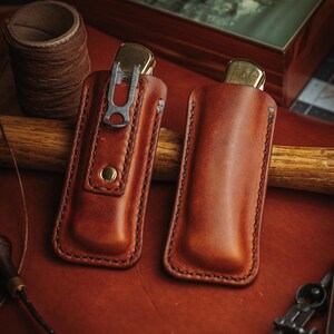 May include: Two tan leather knife sheaths with visible stitching along the edges. One sheath features a metal pocket clip and a brass snap button closure, while the other is a plain slip-style pouch. Both contain a folding knife with a brass bolster.