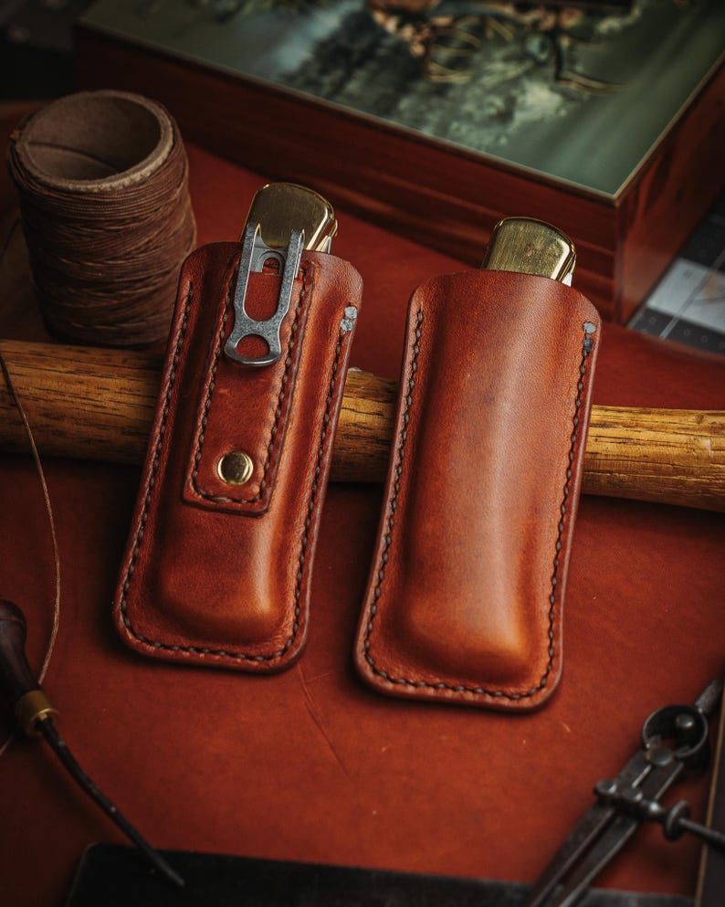 May include: Two tan leather knife sheaths with visible stitching along the edges. One sheath features a metal pocket clip and a brass snap button closure, while the other is a plain slip-style pouch. Both contain a folding knife with a brass bolster.