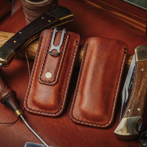 May include: Two rectangular brown leather knife sheaths with visible stitching, one featuring a metal belt clip and a brass snap button. Two folding pocket knives with wooden handles and brass bolsters are positioned alongside the leather sheaths.