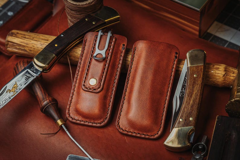 May include: Two rectangular brown leather knife sheaths with visible stitching, one featuring a metal belt clip and a brass snap button. Two folding pocket knives with wooden handles and brass bolsters are positioned alongside the leather sheaths.