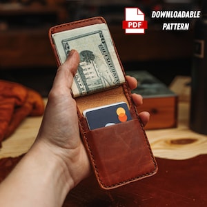 Könnte beinhalten: Ein braunes Lederportemonnaie mit Kartenfach und Geldscheinklammer, das eine Kreditkarte und einen Stapel US-Dollar-Scheine enthält. Das Portemonnaie ist geöffnet und zeigt seinen Inhalt. Der Text "DOWNLOADABLE PATTERN" ist sichtbar.