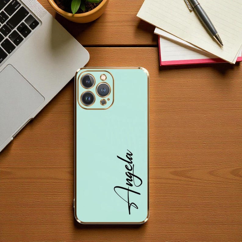 Puede incluir: Una funda de tel&eacute;fono verde menta con un borde dorado. La funda tiene el nombre "Angela" en escritura negra. La funda del tel&eacute;fono est&aacute; sobre una superficie de madera, con un port&aacute;til y un bloc de notas en el fondo.
