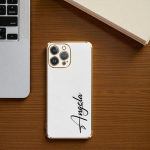 Puede incluir: Funda blanca para iPhone con borde y anillo de c&aacute;mara dorados. El nombre "Angela" est&aacute; escrito en cursiva en la funda. La funda est&aacute; sobre una superficie de madera junto a un port&aacute;til y una pila de papeles.