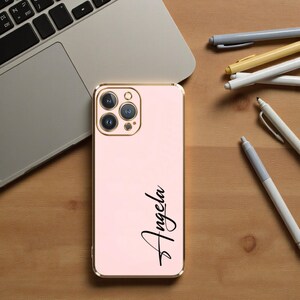 Puede incluir: Una funda de tel&eacute;fono rosa con un marco dorado y el nombre "Angela" en escritura negra. La funda est&aacute; junto a un port&aacute;til y varios bol&iacute;grafos sobre una superficie de madera.