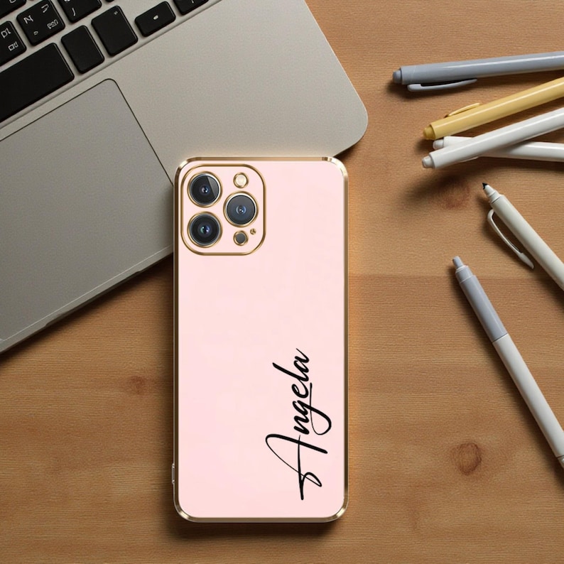 Puede incluir: Una funda de tel&eacute;fono rosa con un marco dorado y el nombre "Angela" en escritura negra. La funda est&aacute; junto a un port&aacute;til y varios bol&iacute;grafos sobre una superficie de madera.