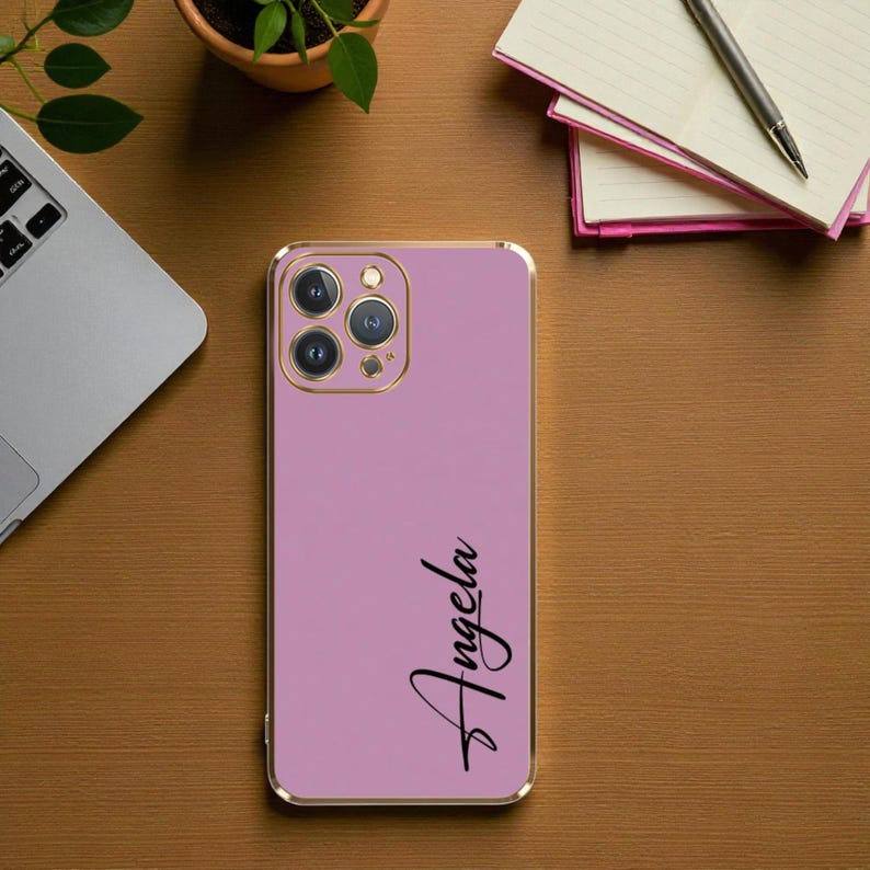 Puede incluir: Una funda de tel&eacute;fono lavanda con un borde dorado. El nombre "Angela" est&aacute; escrito en negro en la funda. La funda del tel&eacute;fono tiene un m&oacute;dulo de c&aacute;mara con tres lentes.