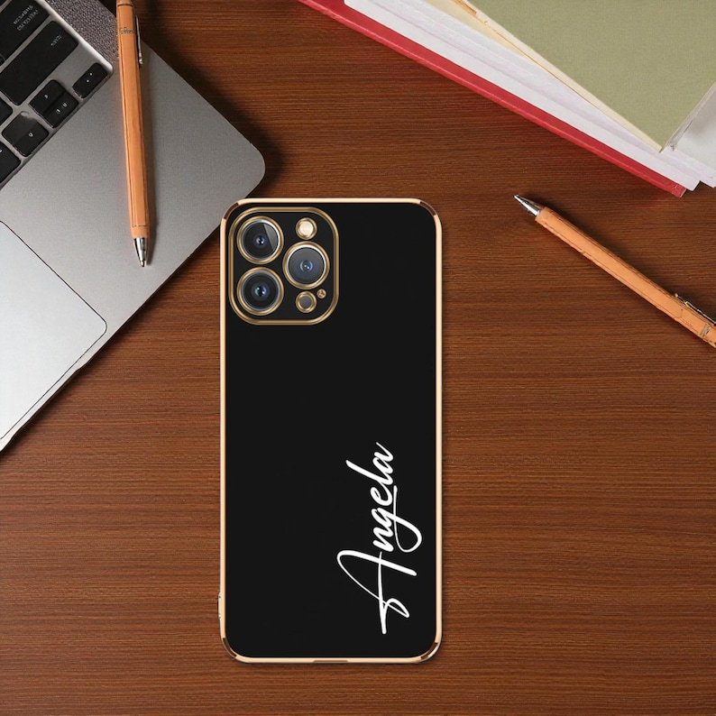 Puede incluir: Funda de tel&eacute;fono negra con borde dorado. La funda tiene el nombre "Angela" escrito en blanco con una fuente cursiva. La funda est&aacute; sobre un escritorio de madera junto a un port&aacute;til y un bol&iacute;grafo.