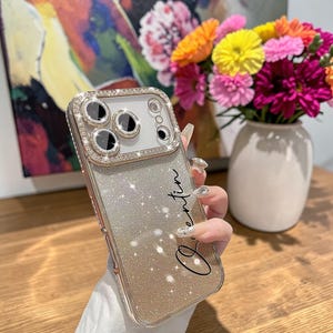 May include: A gold and silver glitter phone case with a diamond-encrusted camera lens frame. The case has the name "Quentin" written in cursive. A bouquet of colorful flowers sits in a white vase in the background.