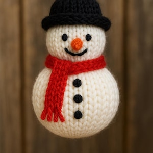 May include: A handmade snowman ornament with a black hat, black button eyes, and a carrot nose. The snowman wears a red scarf and has three black buttons. The ornament is made of knitted material.
