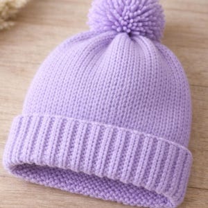 Könnte beinhalten: Ein lilafarbener Strickmütze mit passendem Bommel oben. Die Mütze hat einen gerippten Bund und ein weiches, strukturiertes Aussehen. Dieses Winteraccessoire ist ideal für kaltes Wetter.