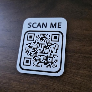 May include: A rectangular white sign with rounded corners featuring a black QR code in the center and the text SCAN ME printed in bold black capital letters above the code. The sign has a thin black border surrounding the QR code graphic.