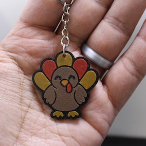 May include: A turkey keychain featuring a brown body, a red wattle, and a tail with alternating red and gold feathers. The design is outlined in black and includes a metal chain and ring attachment for securing keys or other small items.
