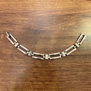 May include: A silver-toned bracelet with a geometric design. The bracelet features rectangular links connected by smaller, rectangular bars. The links are arranged in a curved shape, and the bracelet is displayed on a wooden surface.
