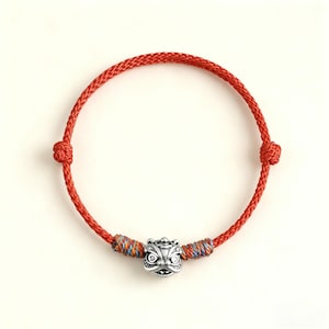 Peut inclure: Bracelet tressé rouge avec un charm en forme de tête de lion argenté. Le bracelet comporte des nœuds réglables et des accents colorés près du charm. Un bijou simple mais saisissant.