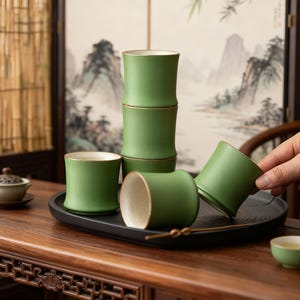 Può includere: Un set di sei tazze da tè in ceramica verde con un design a forma di bambù. Le tazze hanno una forma cilindrica con bordo dorato e interno chiaro. Sono disposte su un vassoio nero su un tavolo di legno.