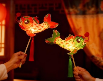 May include: Two colorful fish-shaped paper lanterns, held by hands, are illuminated. The lanterns feature red, green, and yellow scales, with red tassels and wooden handles. The background is a dark, warm-toned interior.
