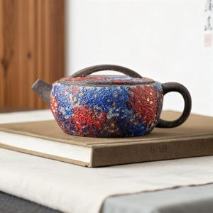 Puede incluir: Pequeña tetera de cerámica con un diseño moteado en rojo, azul y dorado. La tetera tiene un asa y un pico de color marrón oscuro y está sobre un libro cerrado. La tetera probablemente se usa para preparar té.