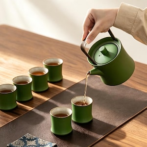 May include: A matte green ceramic teapot with a dark brown handle pouring tea into a matching green ceramic cup. The set includes five additional green ceramic cups with light-coloured rims, all featuring a smooth, cylindrical, slightly flared design.