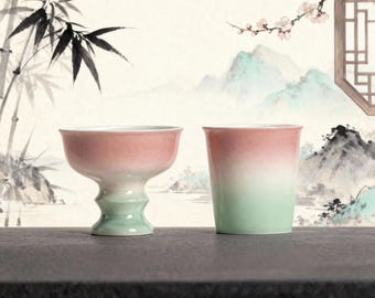 May include: Two ceramic teacups with a gradient color scheme of pink and green. One cup is a footed bowl, and the other is a straight-sided tumbler. The background features a traditional Asian-style painting with bamboo and mountains.