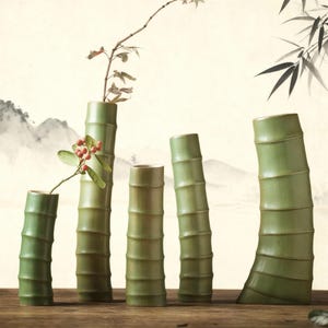 May include: Five green ceramic vases, shaped like bamboo, of varying heights. Some hold small branches with leaves and berries. The background shows a painted landscape. The vases are made of ceramic.